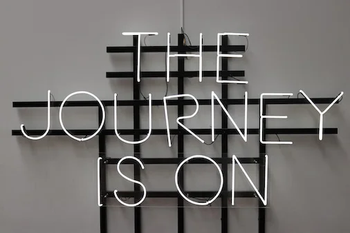 a white neon sign on a gray wall that says 'The Journey Is On'