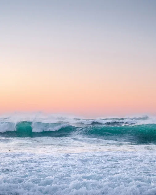 photo of the ocean at sunset with turqoise and white waves and a peach, orange, and light blue sky